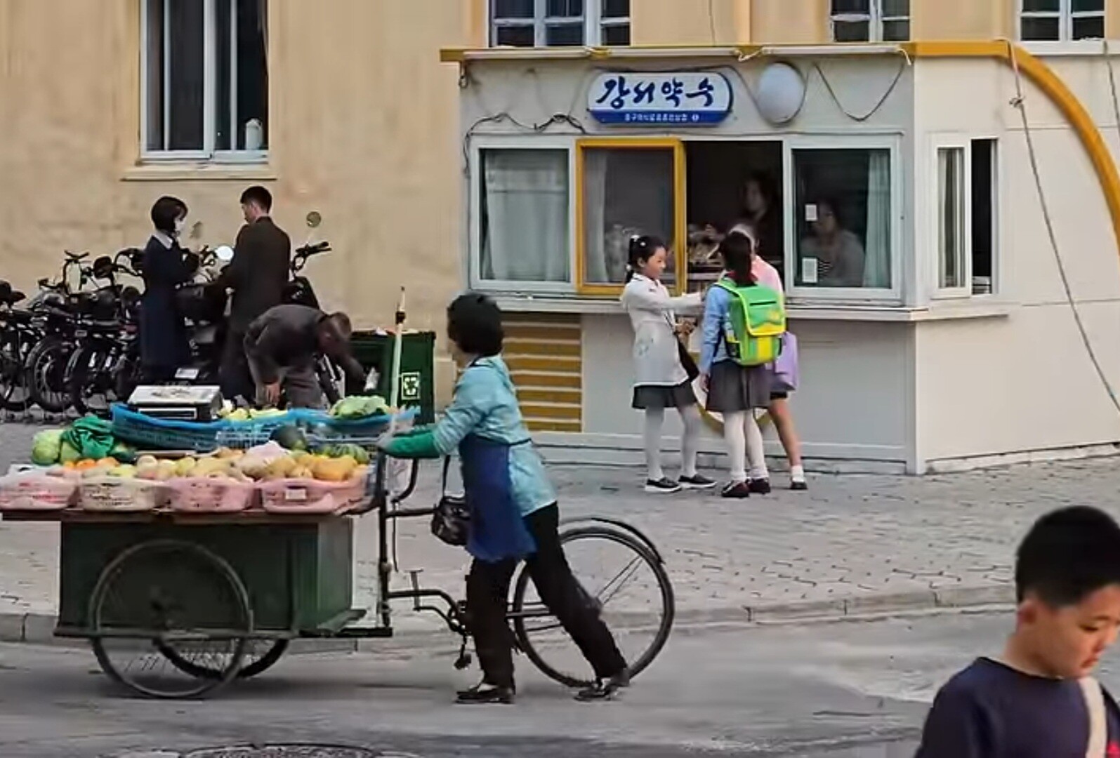 리어카에 과일과 채소를 싣고 가는 북한 상인과 '강서약수'라 적힌 상점 앞에 서 있는 학생들. 사진=유튜브 캡처